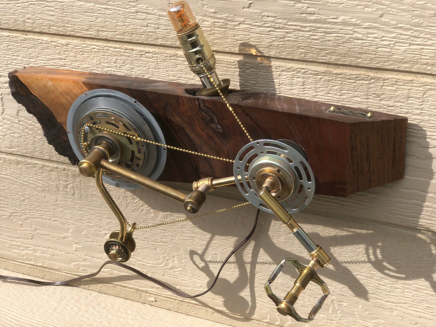 Three-quarter view of Metal Gear Solid with the Edison filament bulb glowing at the top of the wood slab. Two metal disc gears and a brass crank-and-pedal mechanism are visible, connected by a bead chain that runs from the pedal up to the bulb socket. A wall-mounted, pedal-actuated lamp made from antique lamp parts on hand-carved reclaimed wood, by Random Brass