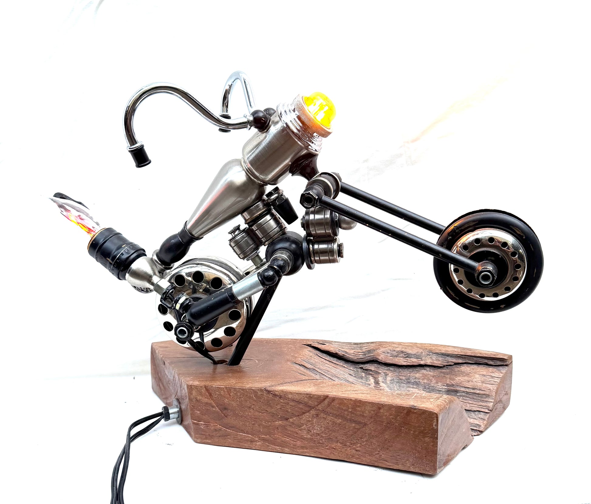 Chopping Block, a sculptural chopper-style motorcycle lamp built from antique chrome and steel lamp parts on a hand-carved reclaimed wood base. The piece features an extended front fork, raked-out front wheel, curved chrome handlebars, blacked-out wheels with chrome hubs, and a small amber Edison filament bulb mounted at the headlight position. A fully functional lamp by Random Brass.