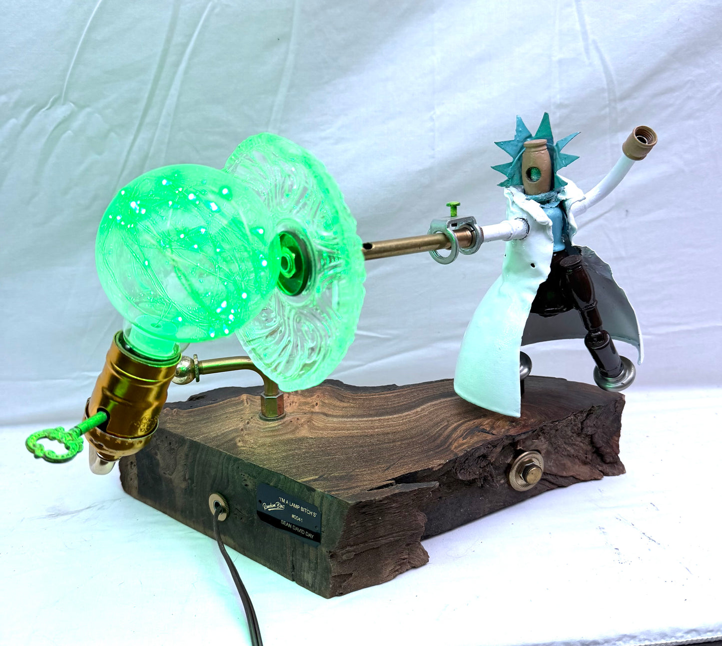 I'm a Lamp, Bitches!, a Rick and Morty-inspired sculptural lamp by Random Brass. A figure of Rick Sanchez with blue spiked hair, white lab coat, and one arm raised stands on a live-edge reclaimed walnut base. A large green glowing portal-style bulb is mounted on a brass socket opposite the figure, casting the scene in green light. A fully functional lamp built primarily from antique lamp parts with sculpted detail.