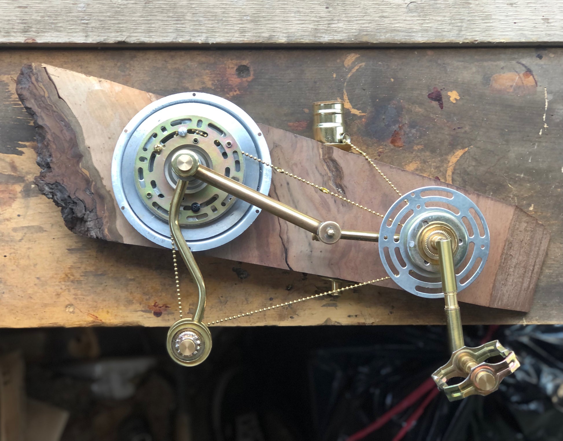 Top-down view of a brass and metal gear-driven lamp mechanism mounted to a live-edge wood slab. Two large disc gears are connected by a brass crank arm and bead chain, with a brass bicycle pedal at the bottom right. From Metal Gear Solid, a pedal-actuated wall-mounted lamp by Random Brass made entirely from antique lamp parts.