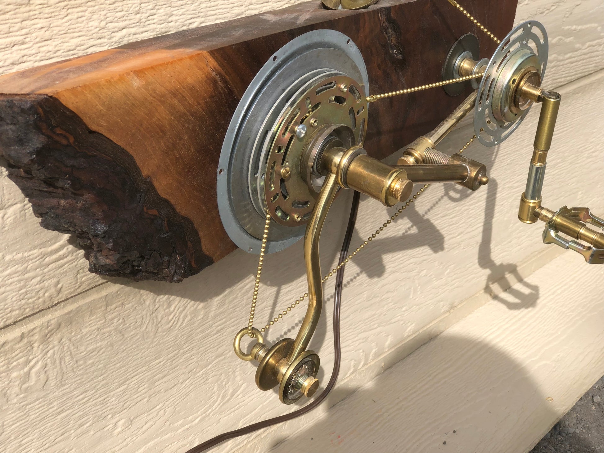Angled view of Metal Gear Solid mounted to a charred live-edge reclaimed wood slab. Two metal gears mounted to the wood are connected by brass crank arms and a bead chain running down to a brass bicycle pedal hanging below the wood. A wall-mounted lamp built entirely from antique lamp parts by Random Brass.