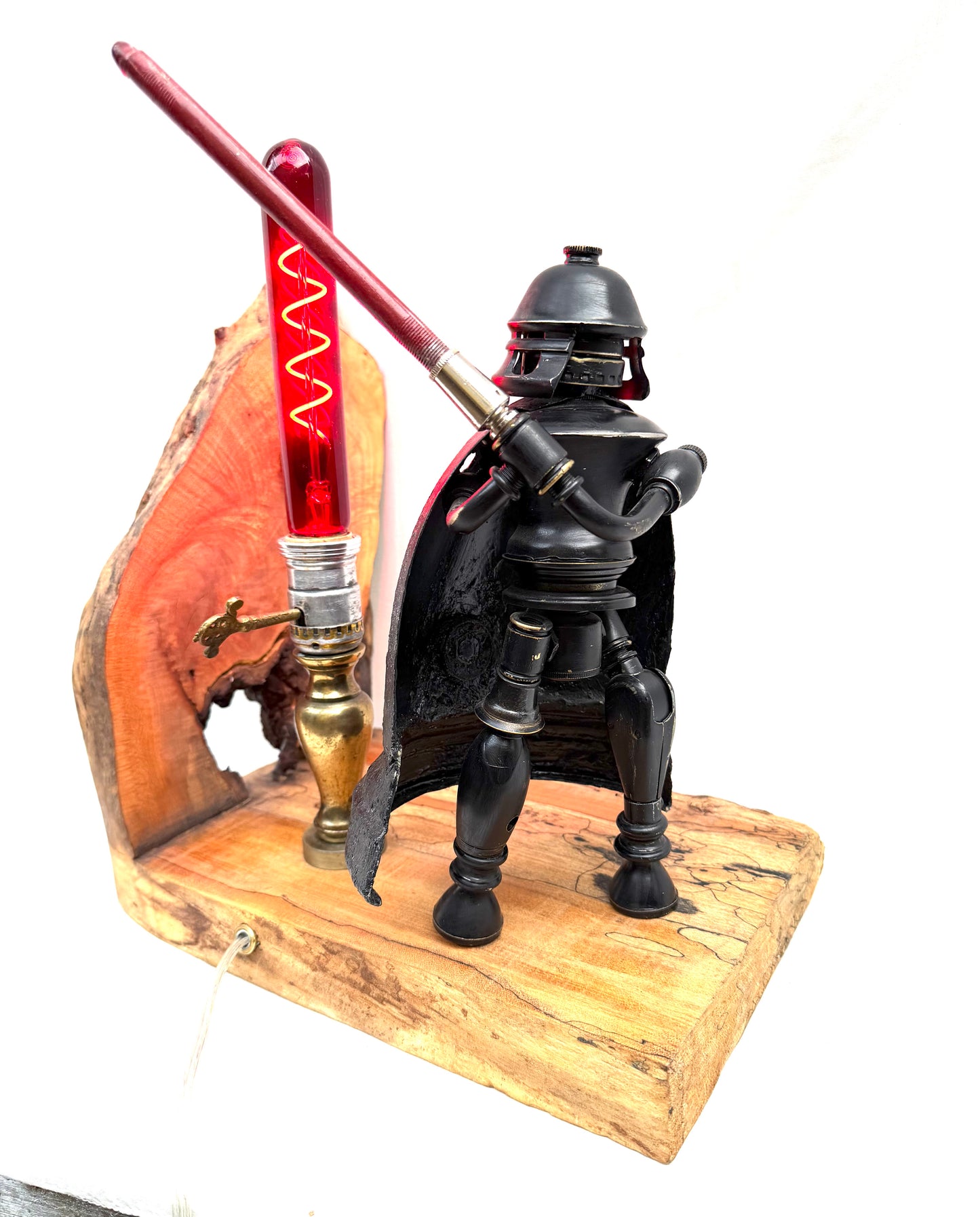 Darth photographed against a clean white background, showing the full sculptural figure built from antique lamp parts. The matte black body, dark cape, and domed helmet stand on a pale spalted reclaimed wood base alongside a glowing red spiral filament tube that serves as the lamp's light source. All natural finishes — nothing painted. A fully functional lamp by Random Brass.