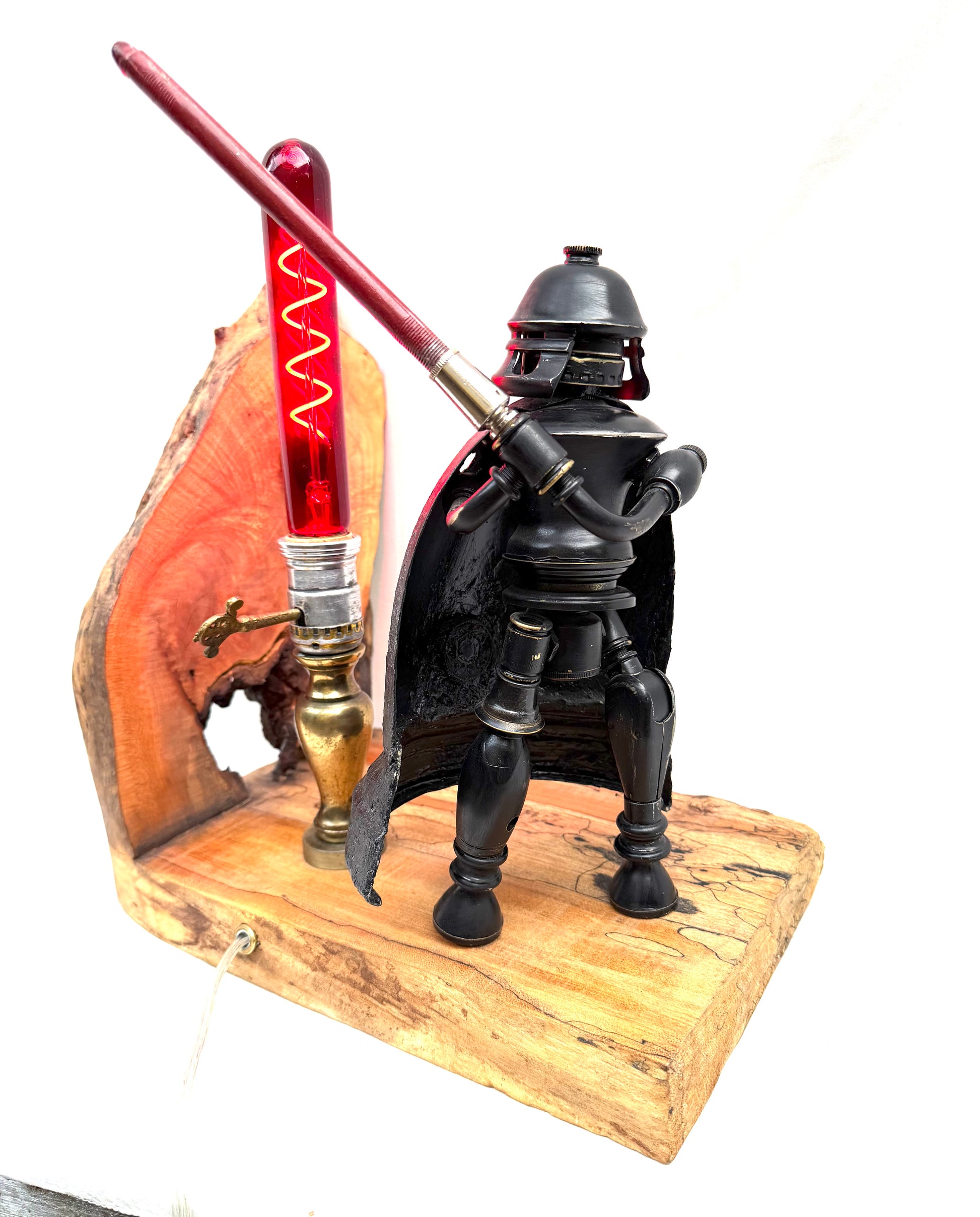 Darth photographed against a clean white background, showing the full sculptural figure built from antique lamp parts. The matte black body, dark cape, and domed helmet stand on a pale spalted reclaimed wood base alongside a glowing red spiral filament tube that serves as the lamp's light source. All natural finishes — nothing painted. A fully functional lamp by Random Brass.