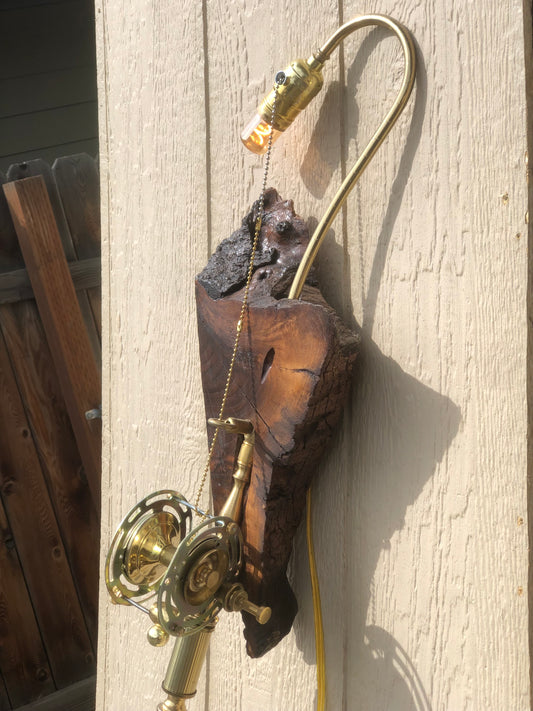 Marlin, a wall-mounted sculptural fishing rod lamp built from antique brass lamp parts on a charred reclaimed wood slab. A long curved brass gooseneck arm extends upward from the wood to a socket and Edison filament bulb at the top, with a beaded pull chain running down to a brass reel mechanism at the bottom. A fully functional lamp by Random Brass.