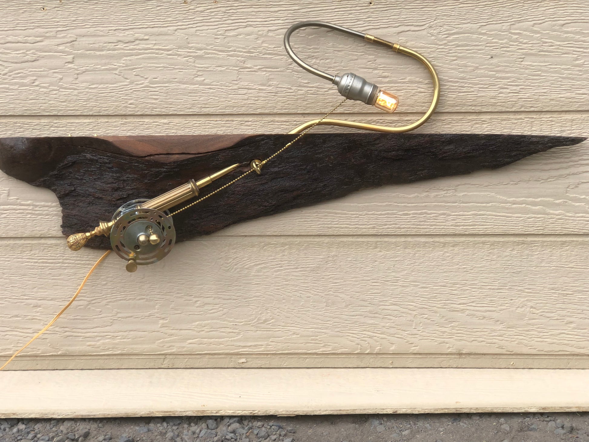 River Spey, a wall-mounted sculptural fly fishing rod lamp built from antique brass lamp parts on a long, charred, live-edge reclaimed wood slab. The slab runs horizontally across the wall with a brass reel and rod at one end, and a curved silver and brass gooseneck arm extending up from the slab to an Edison filament bulb at the top. A fully functional lamp by Random Brass.