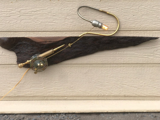 River Spey, a wall-mounted sculptural fly fishing rod lamp built from antique brass lamp parts on a long, charred, live-edge reclaimed wood slab. The slab runs horizontally across the wall with a brass reel and rod at one end, and a curved silver and brass gooseneck arm extending up from the slab to an Edison filament bulb at the top. A fully functional lamp by Random Brass.