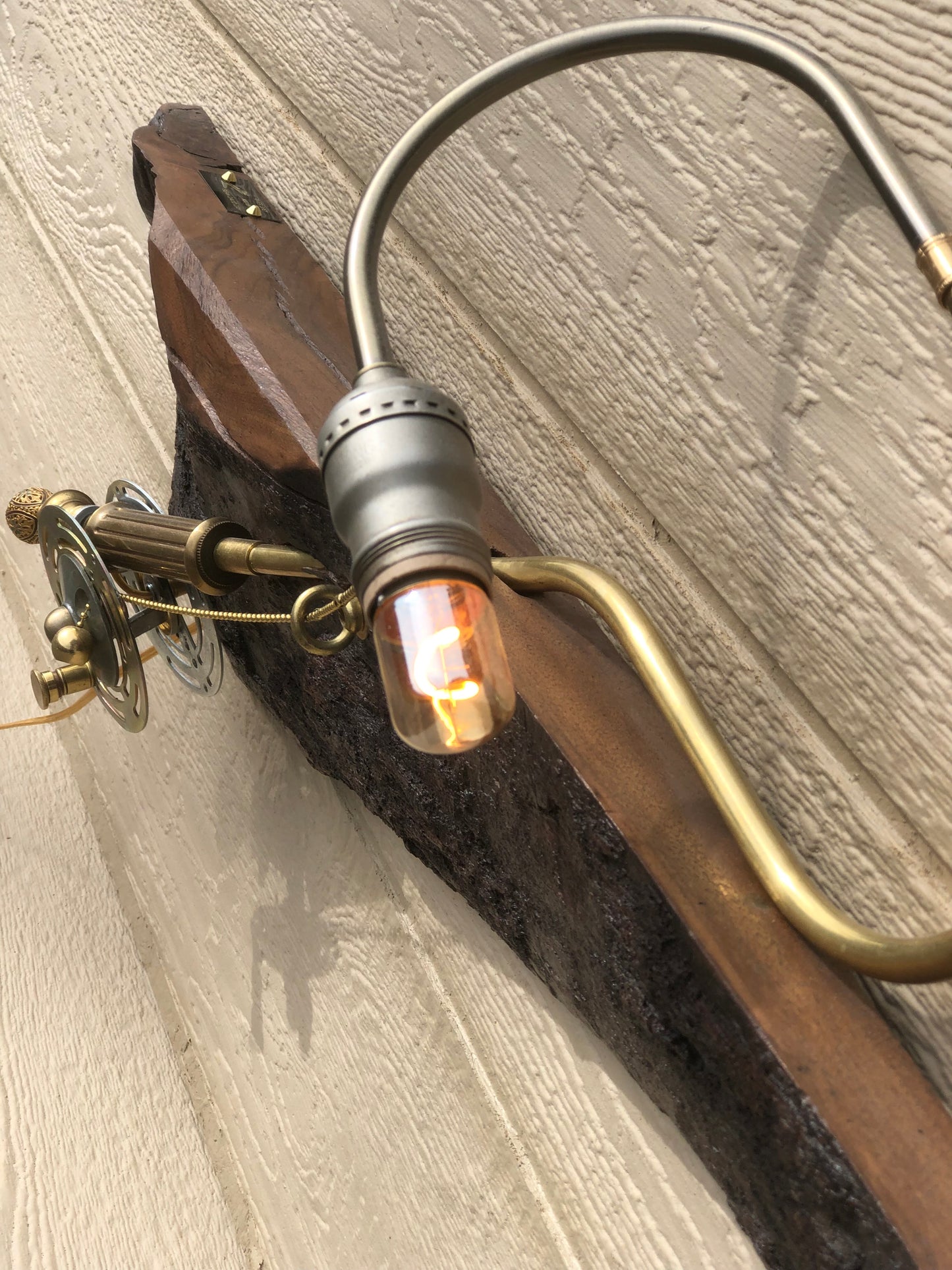 Close-up of River Spey lit, showing the warm glow of the Edison filament bulb at the end of the curved silver and brass gooseneck arm. The dark charred edge of the live-edge reclaimed wood slab is visible alongside the arm, with the brass reel mechanism partially visible in the foreground. A sculptural fly fishing rod lamp by Random Brass.