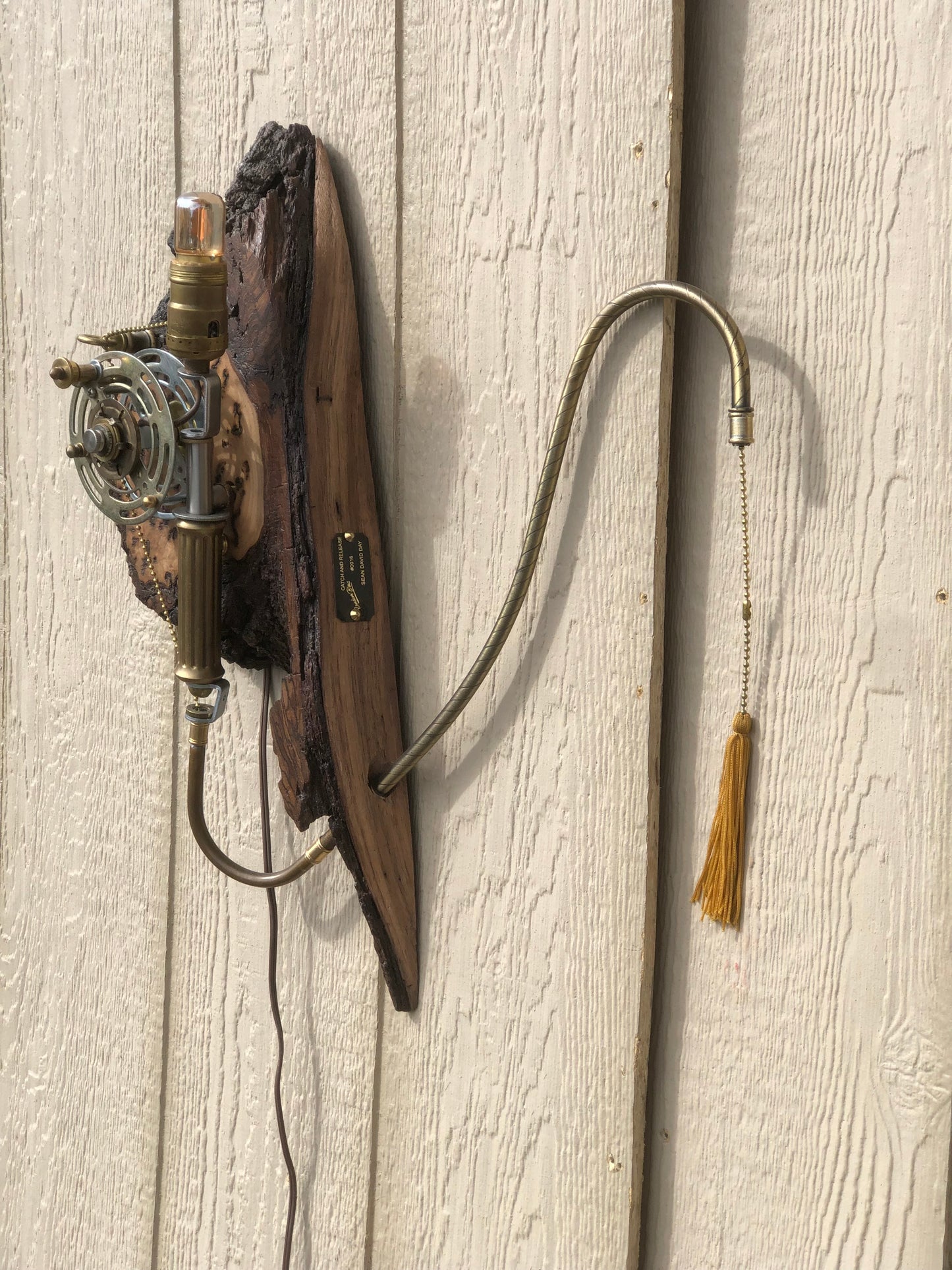Side view of Catch and Release mounted on a wall, showing the full silhouette — burl wood base, brass and silver reel mechanism, Edison filament bulb at the top, and the long gooseneck pull chain extending outward with a gold tassel at the end. The first piece in the Random Brass fishing collection.