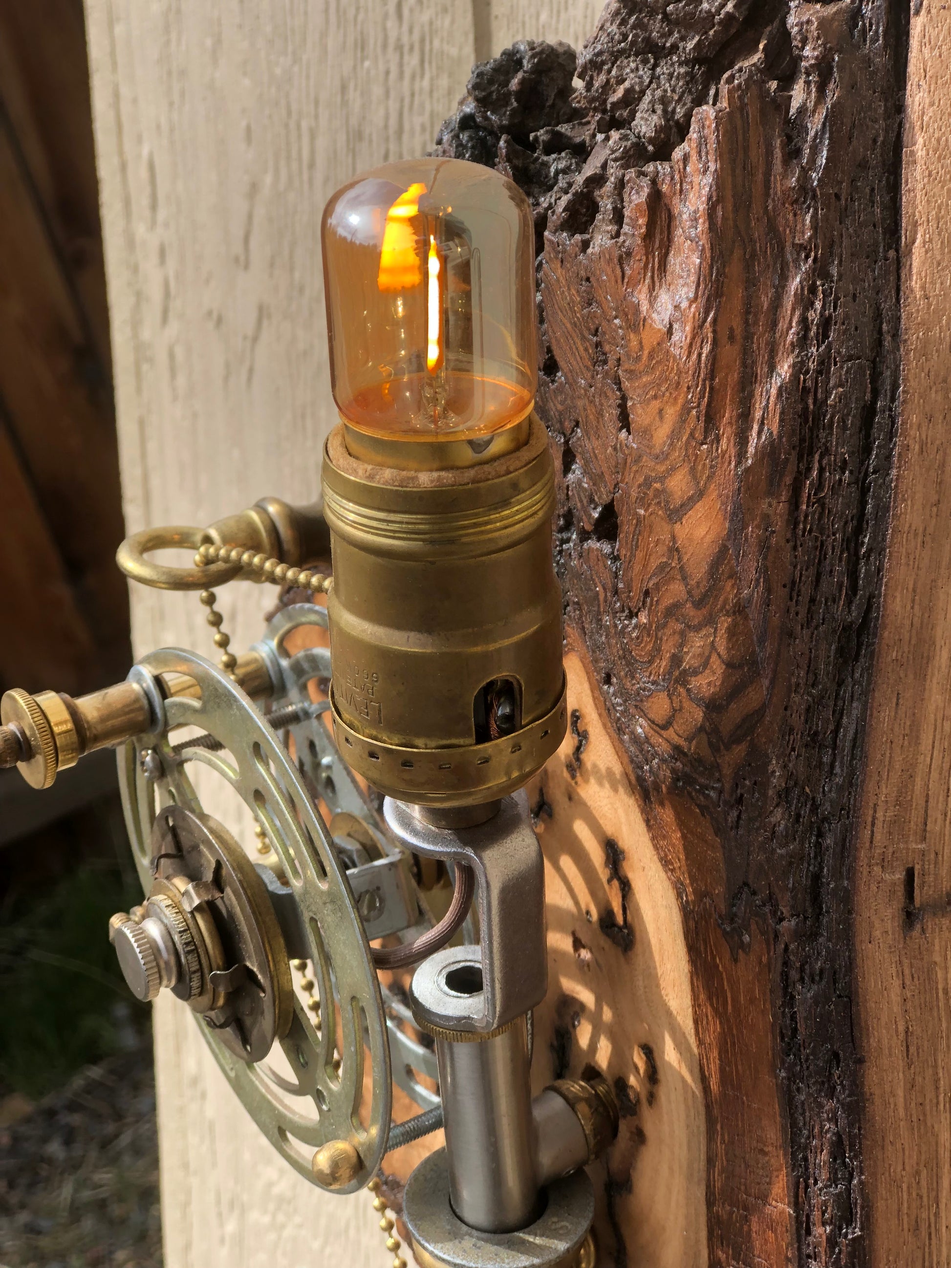 Close-up of Catch and Release lit, showing the warm amber glow of the Edison filament bulb against the dark, deeply textured burl wood base. The brass socket and silver reel hardware are visible in the foreground. A sculptural fishing rod lamp by Random Brass.
