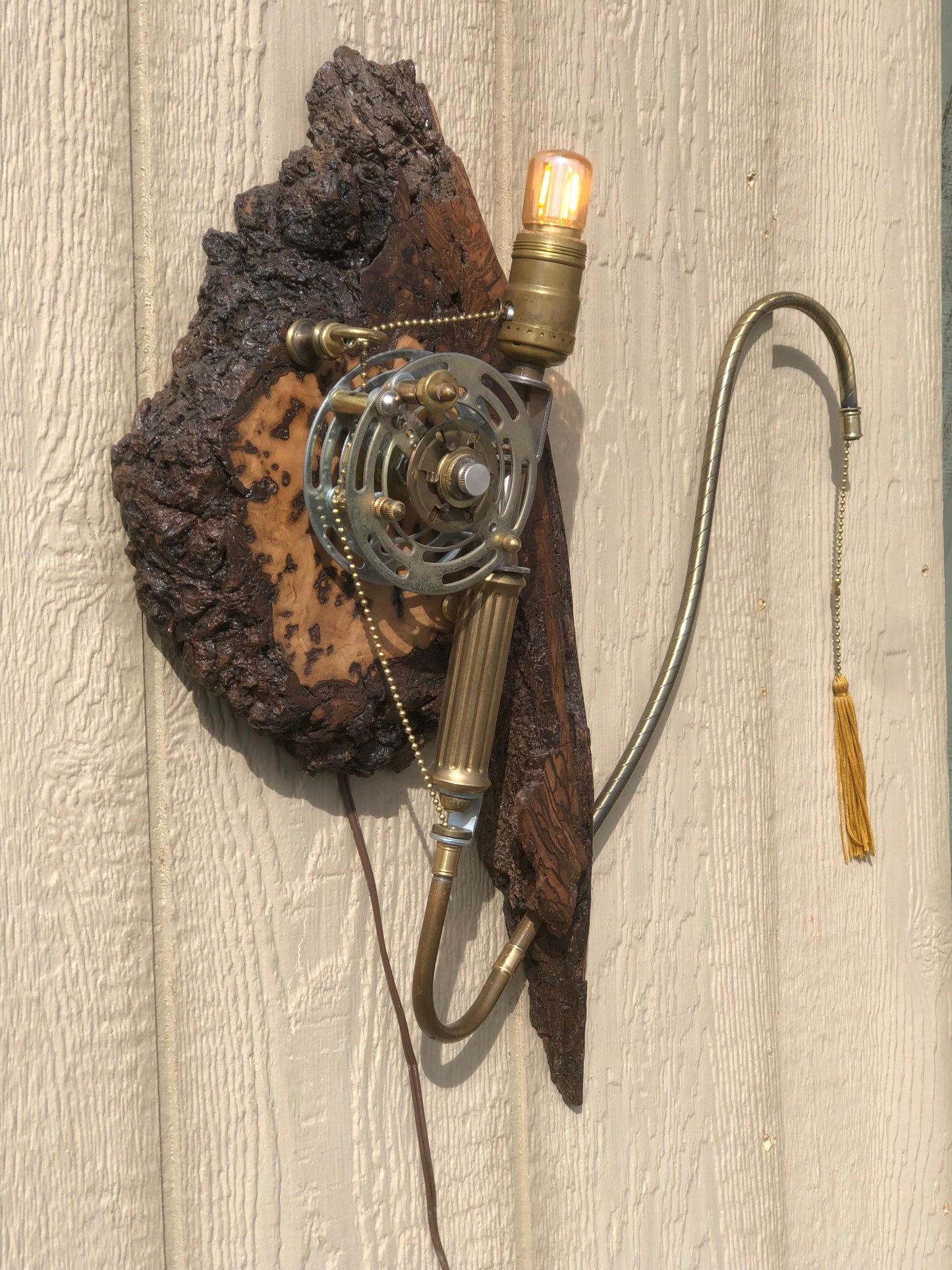 Catch and Release, a wall-mounted sculptural fishing rod lamp built from antique brass and silver lamp parts on a hand-carved burl wood base. A vintage fishing reel mechanism sits at the center, with a beaded line extending out to a gold tassel pull and a small Edison filament bulb glowing at the top. A fully functional lamp by Random Brass.