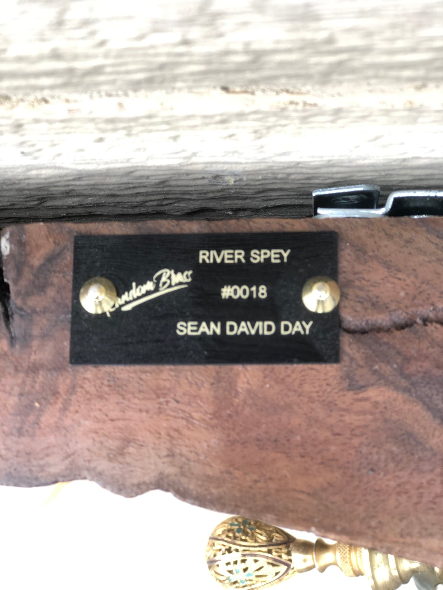 Close-up of the Random Brass nameplate on the back of River Spey. The black plate is engraved in gold with "RIVER SPEY," the piece number "#0018," and the artist's name "SEAN DAY," along with the Random Brass signature logo. Mounted to the reclaimed wood base alongside a decorative brass finial.