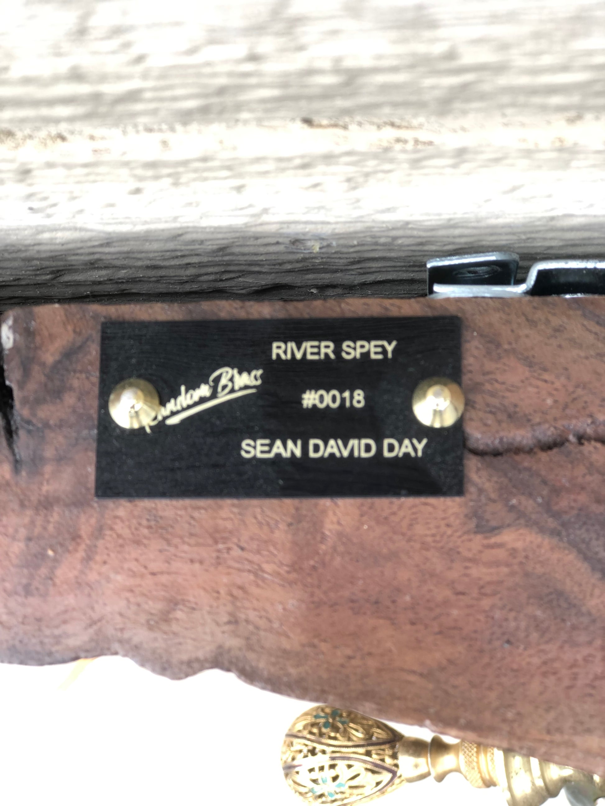 Close-up of the Random Brass nameplate on the back of River Spey. The black plate is engraved in gold with "RIVER SPEY," the piece number "#0018," and the artist's name "SEAN DAY," along with the Random Brass signature logo. Mounted to the reclaimed wood base alongside a decorative brass finial.