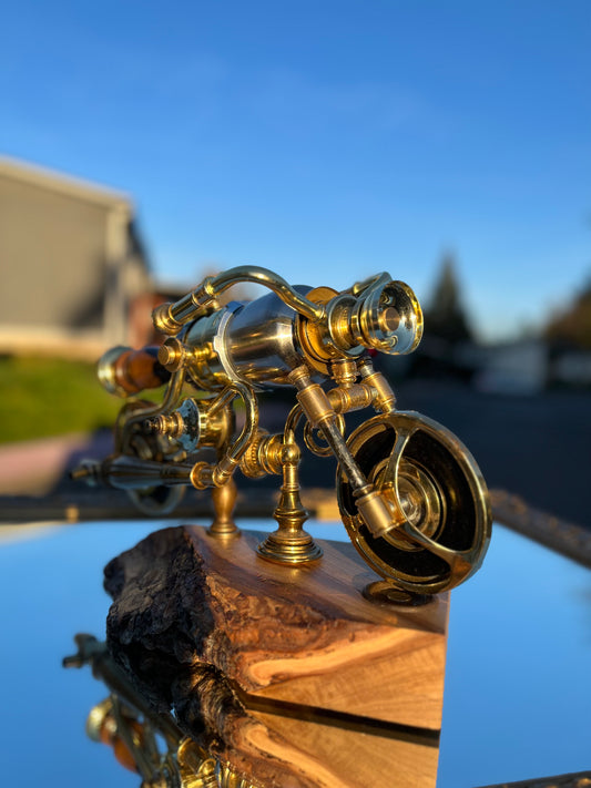 I'm Not Steampunk shown from a three-quarter front angle with the headlamp reflector facing forward. The polished brass body, handlebar arch, and lamp-hardware wheels are visible above a hand-carved live-edge bark base. A sculptural motorcycle lamp built from antique lamp parts by Random Brass.