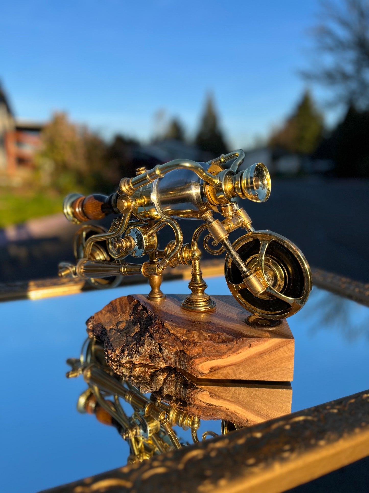 Side profile of I'm Not Steampunk showing the full silhouette of the brass motorcycle — handlebars, fuel tank body, lamp-hardware wheels front and back, and the wood handgrip at the rear. Mounted on a hand-carved live-edge reclaimed wood base. A fully functional lamp by Random Brass.
