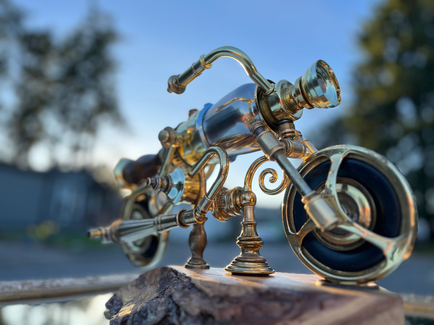 Close-up detail of I'm Not Steampunk highlighting the spiraled brass scrollwork between the handlebars and frame, the curved handlebar arch, and the front wheel built from a brass lamp fitting. Mounted on a hand-carved reclaimed wood base. A sculptural motorcycle lamp by Random Brass.


