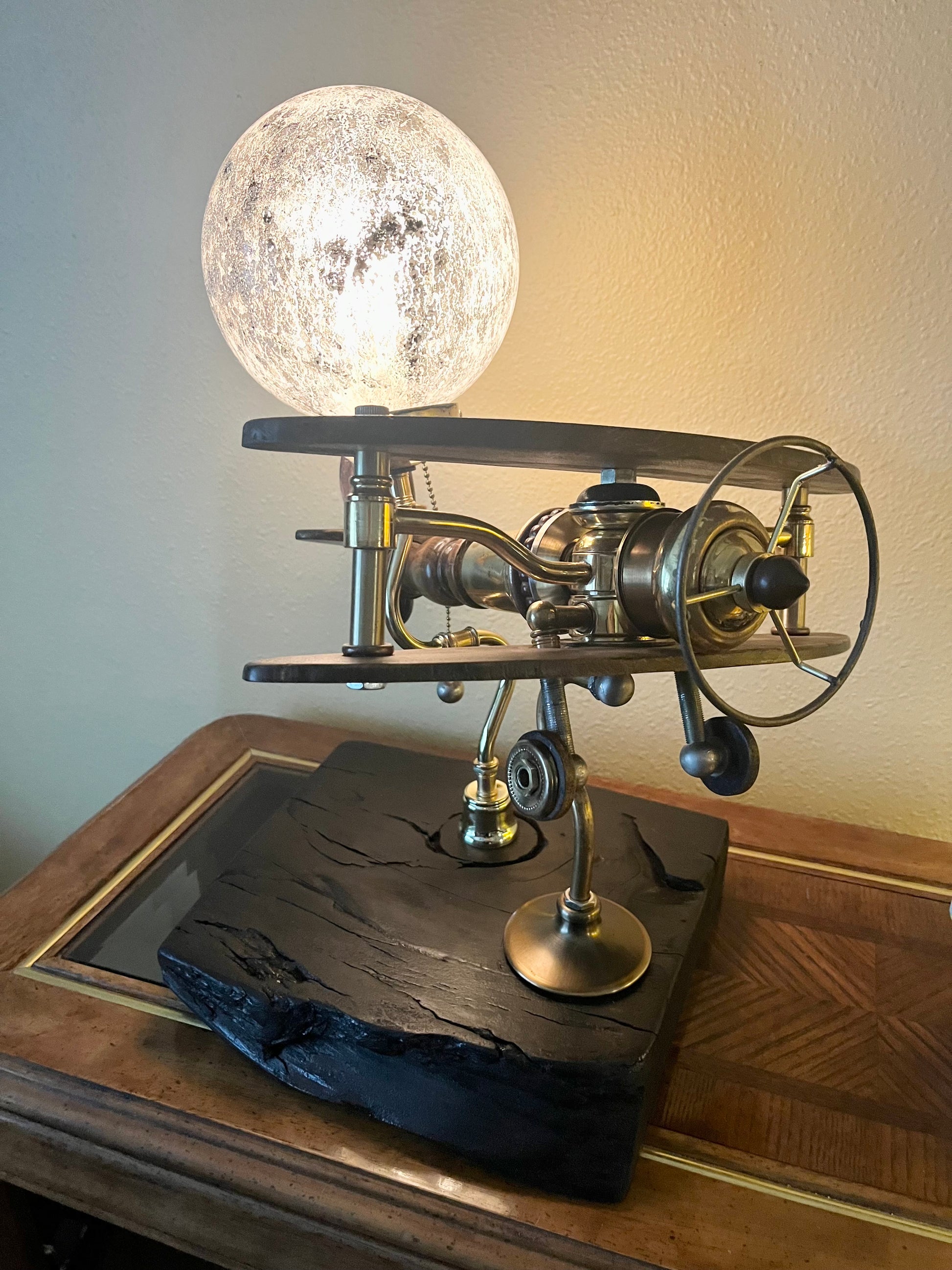 Sinatra, a sculptural biplane lamp with a glowing crackled-glass moon-effect globe mounted above black walnut wings. The brass body sits between the wings with antique lamp hardware forming the propeller, fuselage, and tail. Mounted on a hand-carved charred black reclaimed wood base. A fully functional lamp made from antique lamp parts by Random Brass.