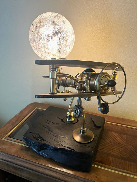 Sinatra, a sculptural biplane lamp with a glowing crackled-glass moon-effect globe mounted above black walnut wings. The brass body sits between the wings with antique lamp hardware forming the propeller, fuselage, and tail. Mounted on a hand-carved charred black reclaimed wood base. A fully functional lamp made from antique lamp parts by Random Brass.