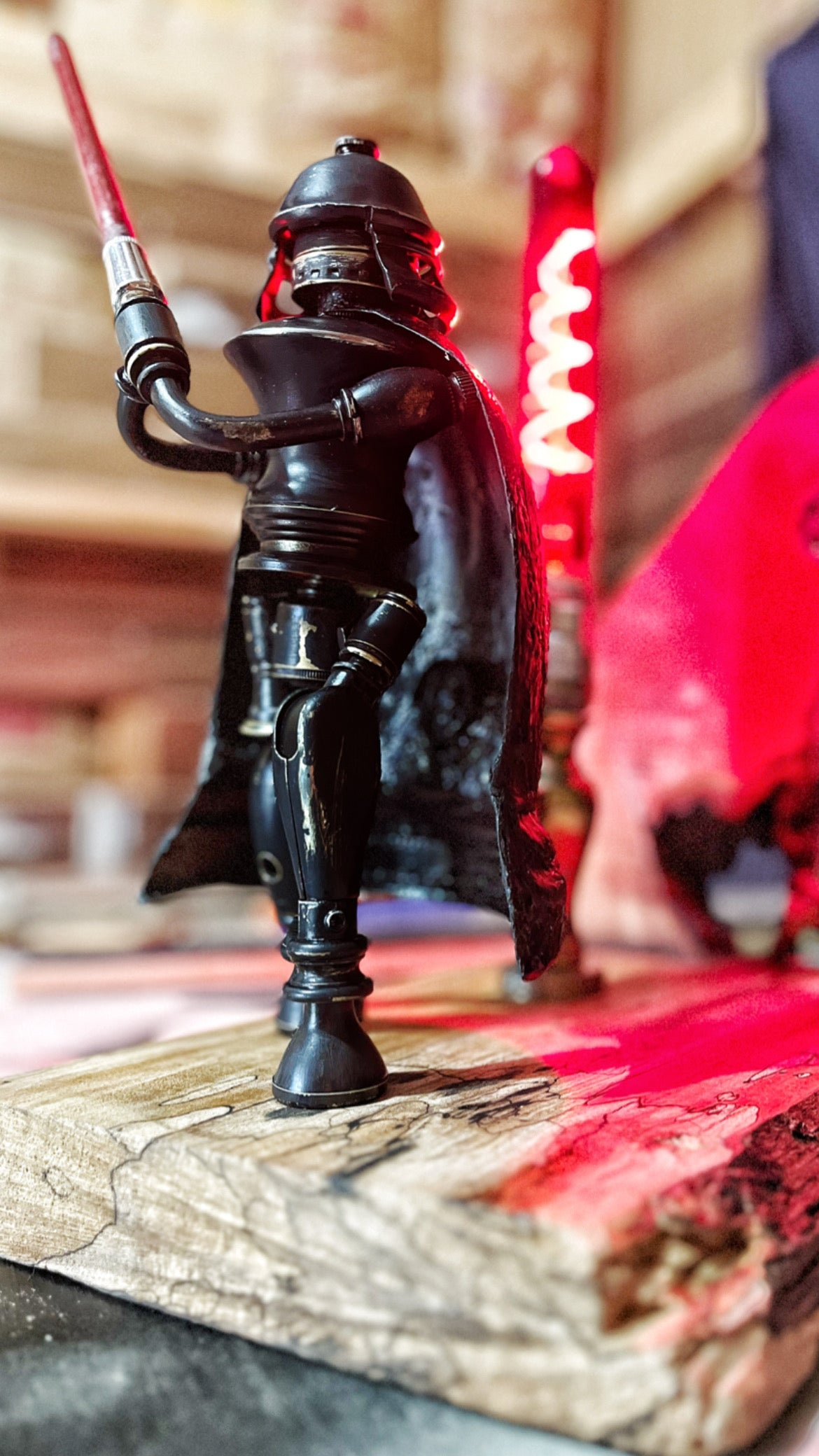 Darth, a sculptural Sith-inspired figure built from antique lamp parts on a hand-carved reclaimed wood base. The matte black figure stands with a sculptural lightsaber held aloft, a flowing red cape behind, and a domed helmet with a visor face. A red filament tube glows beside the figure, casting the surrounding scene in deep crimson. A fully functional lamp by Random Brass.