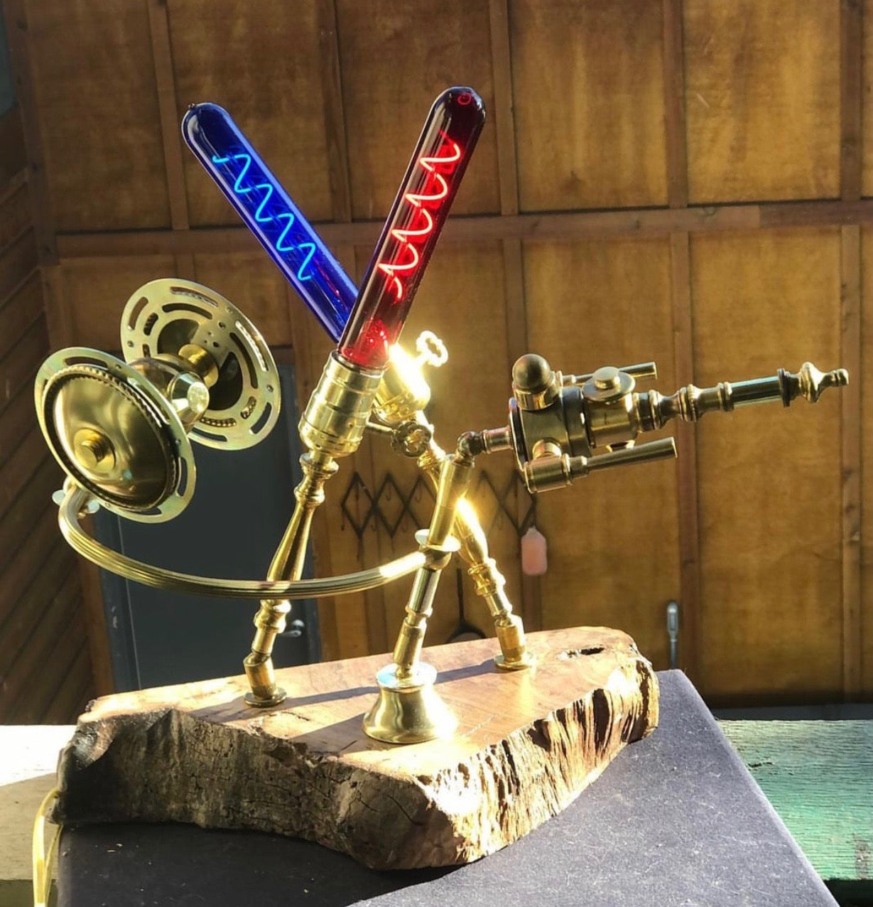 Battle of Endor, a sculptural lamp built from antique brass lamp parts on a live-edge reclaimed wood base. A brass figure with a gyroscope-like shield on one side and an extended brass blaster arm on the other stands at the center, with two crossed glowing filament tubes — one blue, one red — rising above as lightsabers. Both tubes are functional lamps. Made entirely from antique lamp parts by Random Brass.