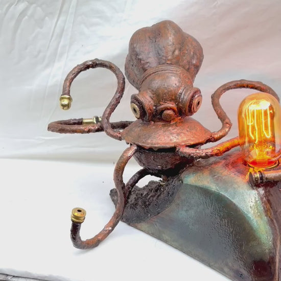 A copper-patinated octopus sculpture with six curling tentacle arms, mounted to a glossy green and teal stone-effect wood base, with a warm Edison filament bulb glowing at the front. Made from antique lamp parts and chandelier components on a hand-carved maple burr base by Random Brass.