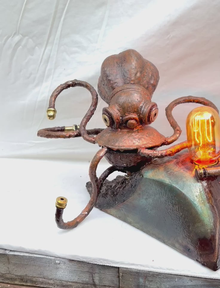 A copper-patinated octopus sculpture with six curling tentacle arms, mounted to a glossy green and teal stone-effect wood base, with a warm Edison filament bulb glowing at the front. Made from antique lamp parts and chandelier components on a hand-carved maple burr base by Random Brass.